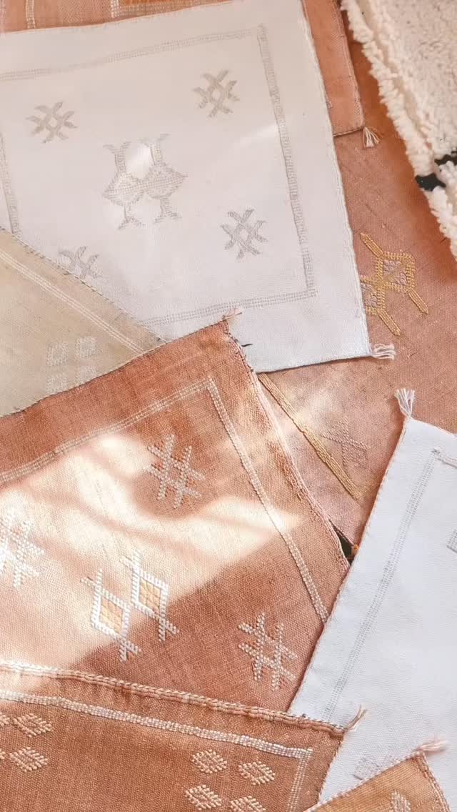 Primer plano de varios textiles cuadrados, posiblemente manteles o tapetes, en tonos terracota, rosa pálido y blanco crudo, con bordados de patrones geométricos y simbólicos bereberes, bañados por luz natural.