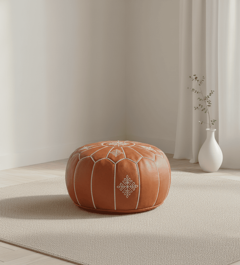 Puf redondo de cuero marrón (58x38 cm) con bordados blancos, en centro de habitación minimalista luminosa, con jarrón blanco y rama, cortinas claras y alfombra beige.