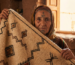Retrato de portada de una anciana mujer amazigh con tatuajes faciales tradicionales sosteniendo una alfombra marroquí artesanal con símbolos geométricos, mostrando la conexión cultural entre la piel y la lana.